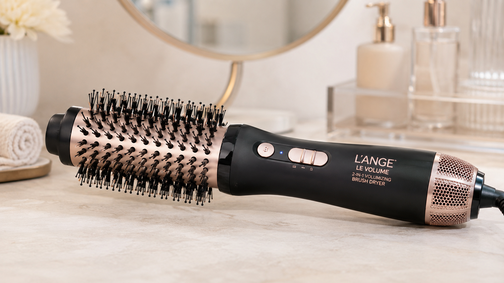 L’ange Le Volume 2-in-1 Volumizing Brush Dryer in black and rose gold displayed on a marble bathroom vanity with soft lighting and luxury beauty styling accessories in the background.