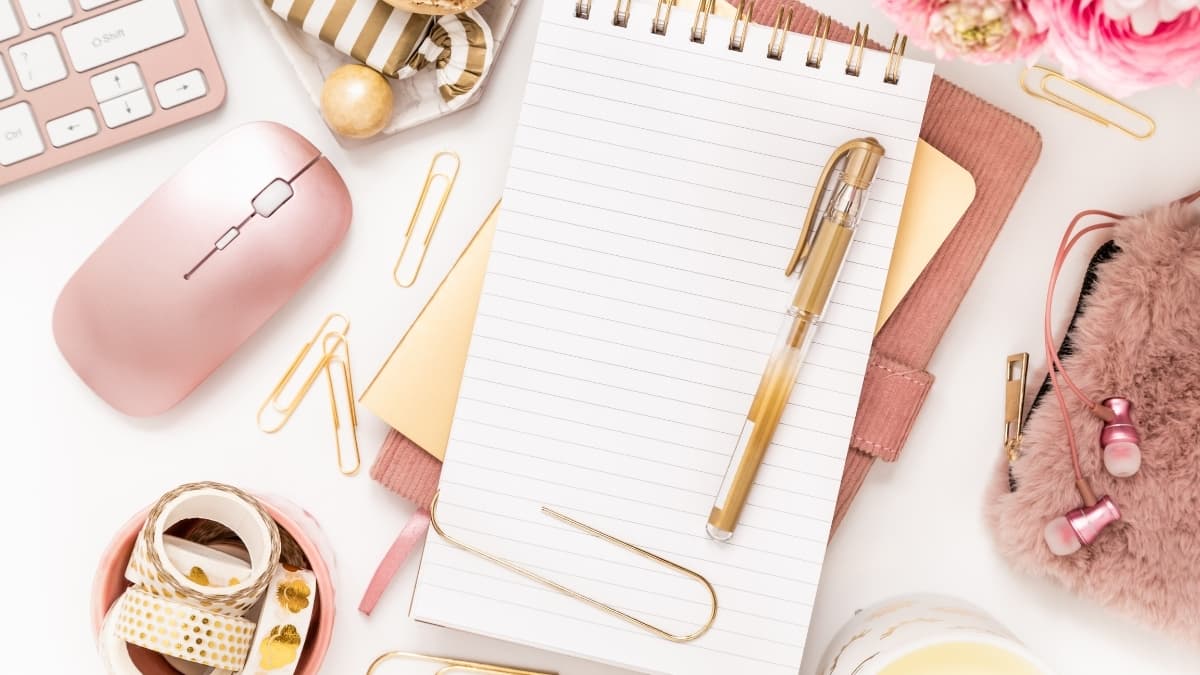 Pink and gold desk accessories with notebook, pen, mouse, paper clips, and earbuds arranged neatly on a stylish feminine workspace desk.