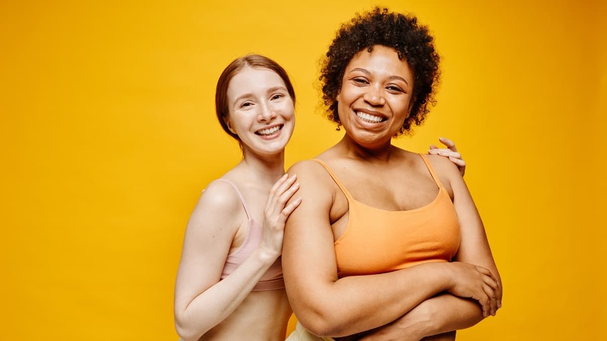 Two women with different body types and skin tones smiling in inclusive wireless bras against a bright yellow background, representing Evelyn & Bobbie’s Inclusivity Promise.