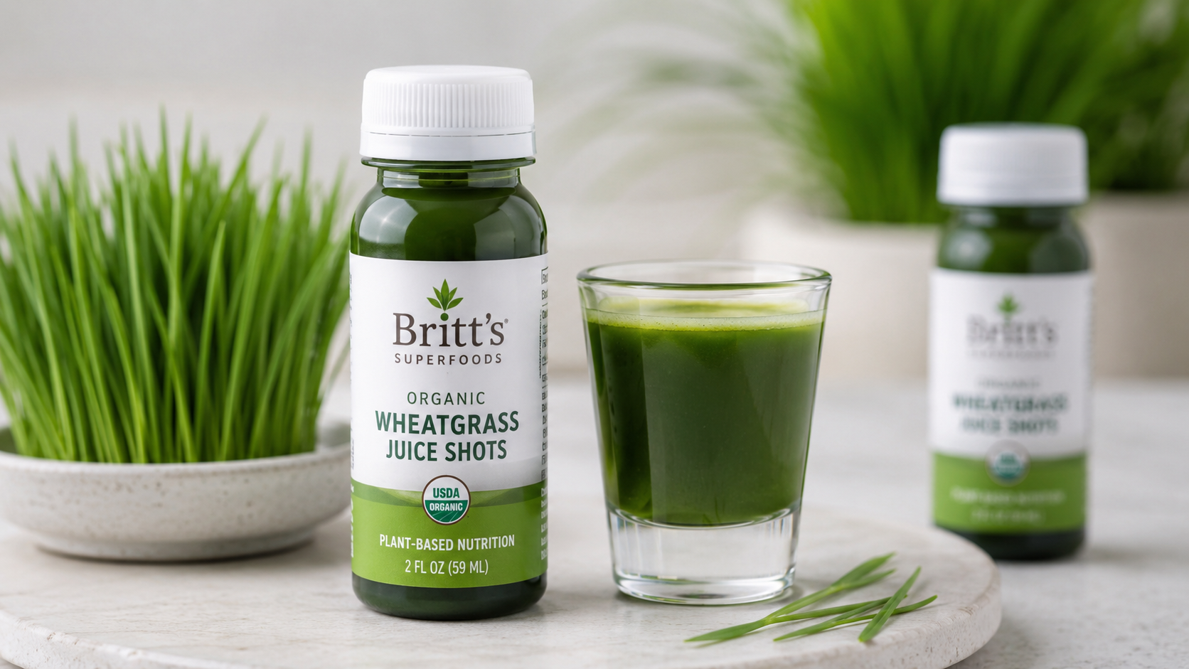 Britt's Superfoods Organic Wheatgrass Juice Shots bottle next to a glass of fresh wheatgrass juice on a white stone tray, surrounded by green wheatgrass plants in a bright natural setting.