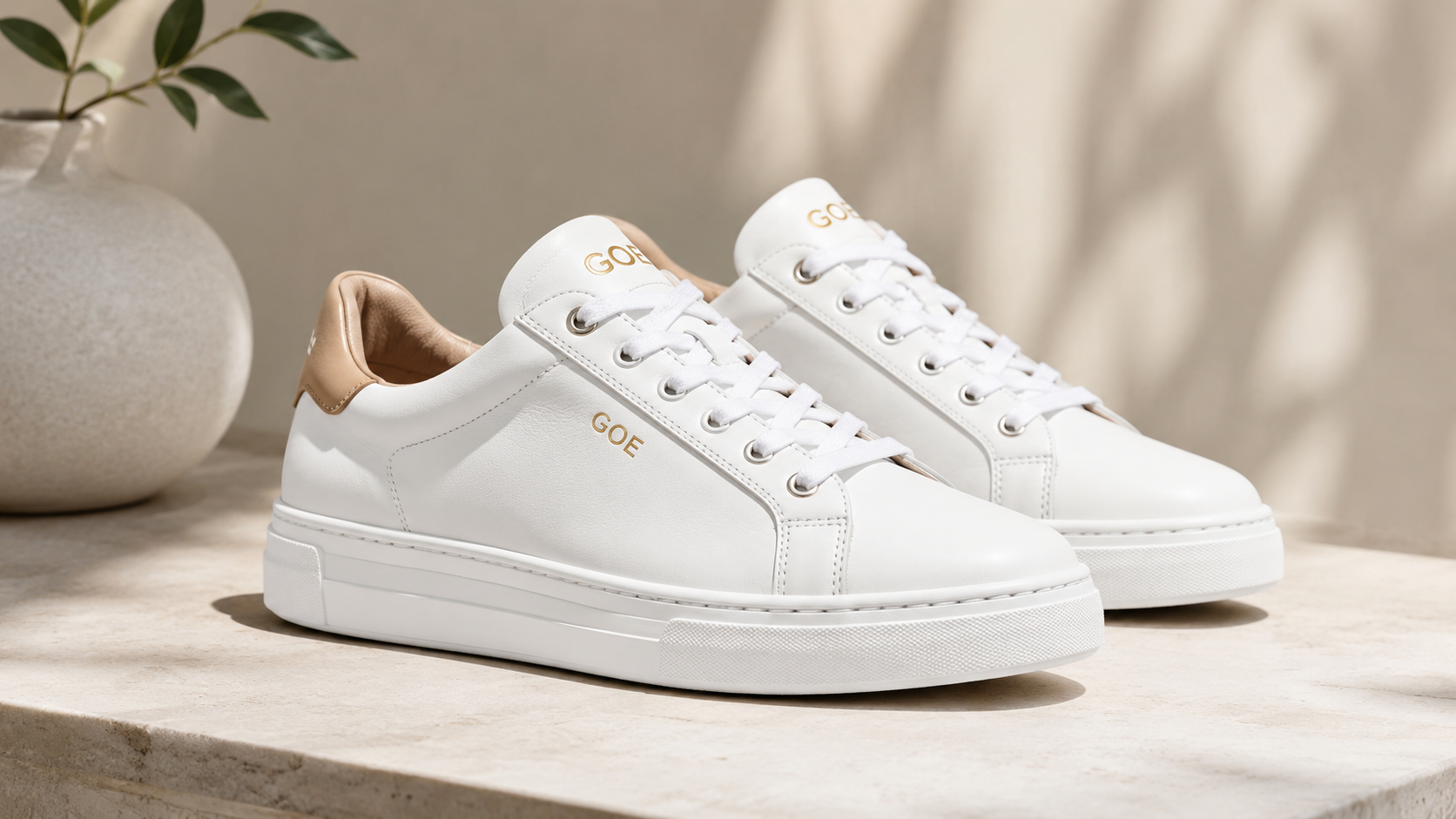 GOE Leather Women's Sneakers in white leather with gold logo accents and beige heel detail, displayed on a stone surface in soft natural light, stylish casual fashion footwear for women.