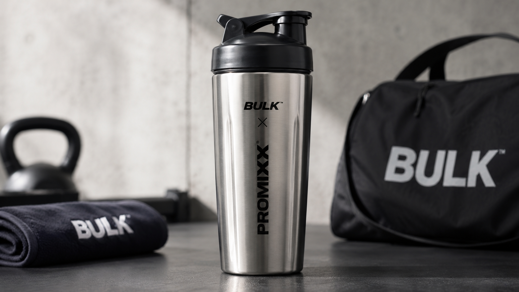 Bulk x Promixx stainless steel protein shaker bottle with black lid on a gym surface, surrounded by fitness accessories in a modern workout setting.