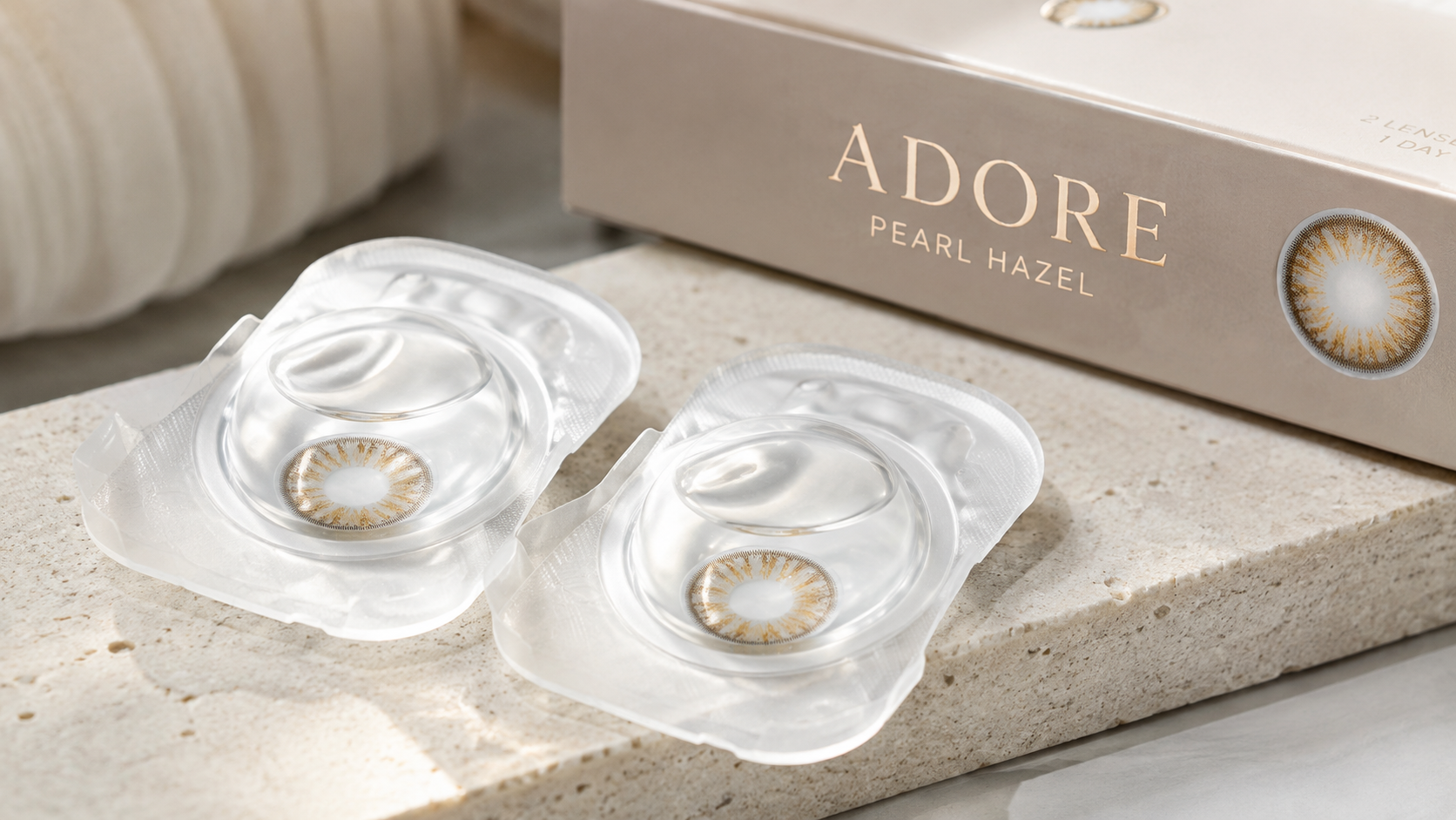 Adore Pearl Hazel colored contact lenses in sealed blister packs beside the Adore Pearl Hazel box on a beige stone surface with soft natural lighting.
